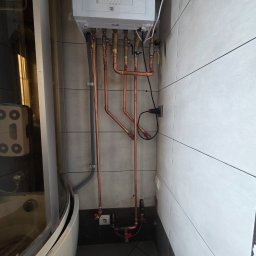 SZOK ARKADIUSZ JEWIARZ - Nowy, biały piec gazowy zamontowany na ścianie z szarymi i białymi płytkami. Widoczne miedziane rury instalacji, podłączenia i elementy armatury. Obok kabina prysznicowa.