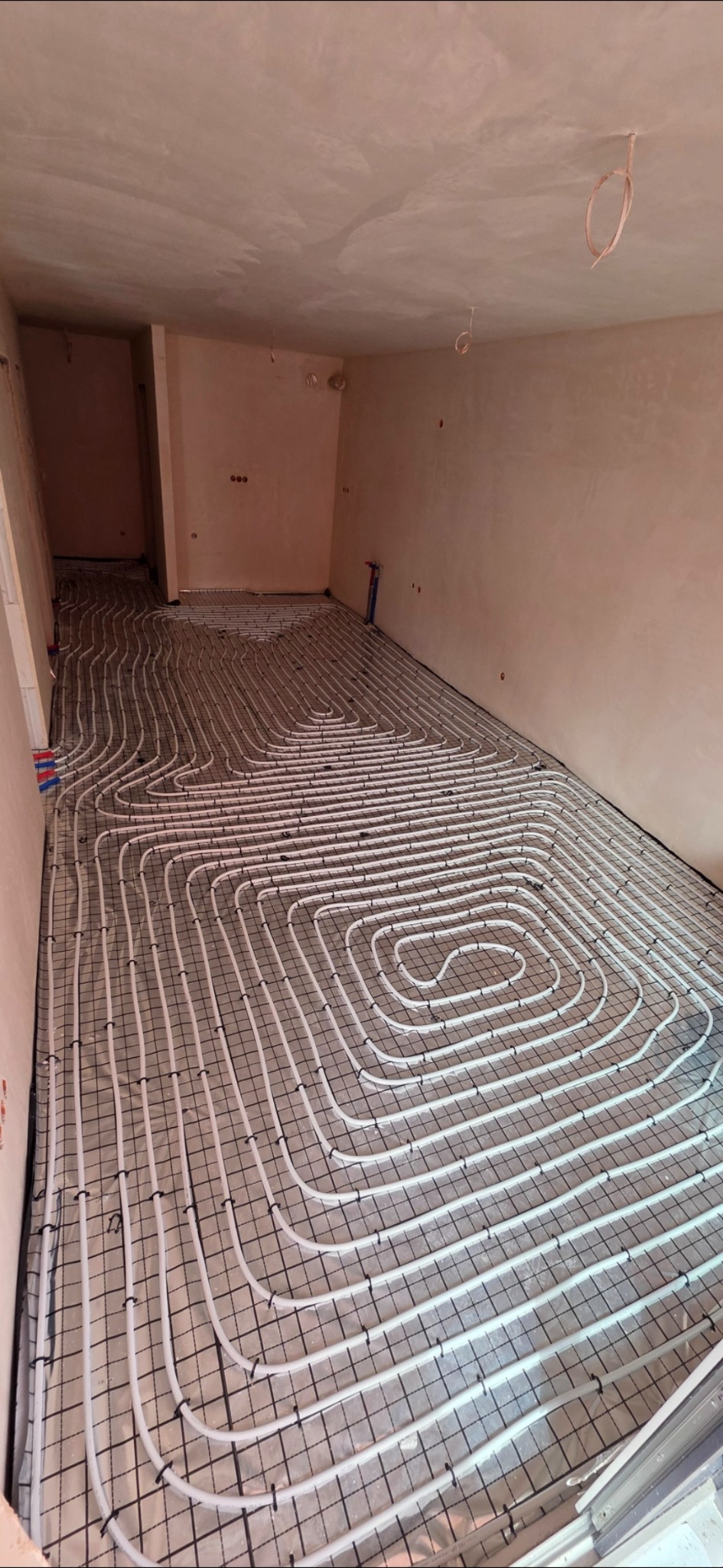 Białe rury ogrzewania podłogowego, ułożone spiralnie na czarnej siatce, w surowym wnętrzu z widocznymi instalacjami elektrycznymi na suficie.