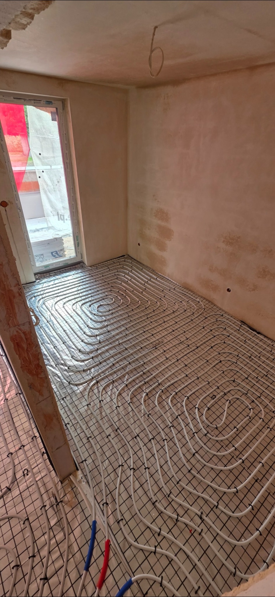 Ułożone rury ogrzewania podłogowego na siatce w surowym wnętrzu z oknem. Elementy instalacji w kolorze niebieskim i czerwonym. Ściany przygotowane do dalszych prac.