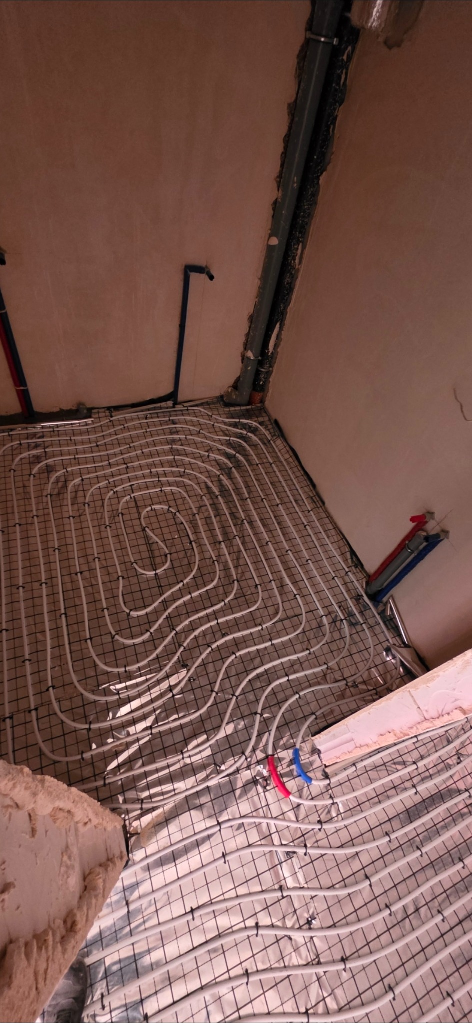 Białe rury ogrzewania podłogowego ułożone spiralnie na czarnej siatce, widoczne elementy instalacji wodnej przy ścianie. Surowy stan wykończenia.