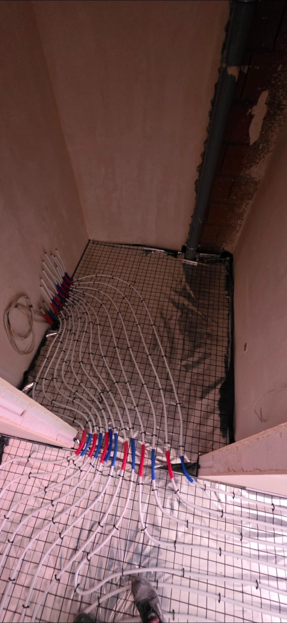 Białe rury ogrzewania podłogowego, z czerwono-niebieskimi oznaczeniami, ułożone na siatce w pomieszczeniu z surowymi ścianami. Widok z góry.