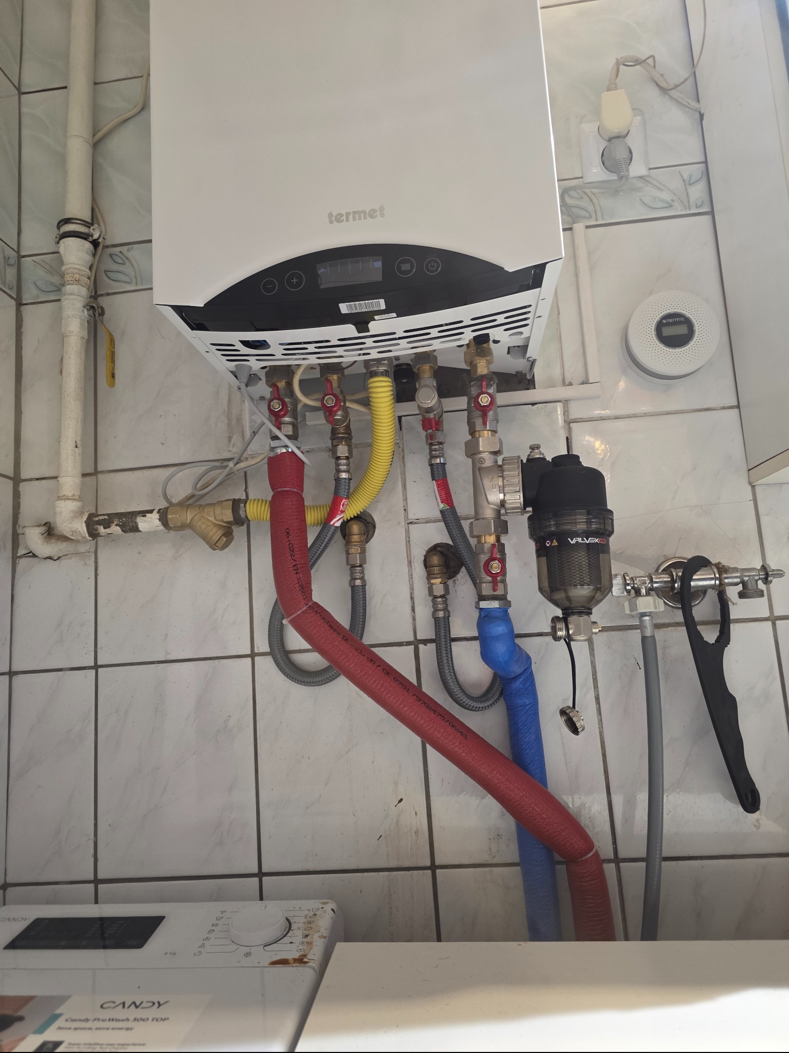 Biały piec gazowy Termet z podłączonymi rurami (czerwona, żółta, niebieska) na tle białych płytek. Widoczny filtr Valvex i detektor gazu Termic. Instalacja w łazience.