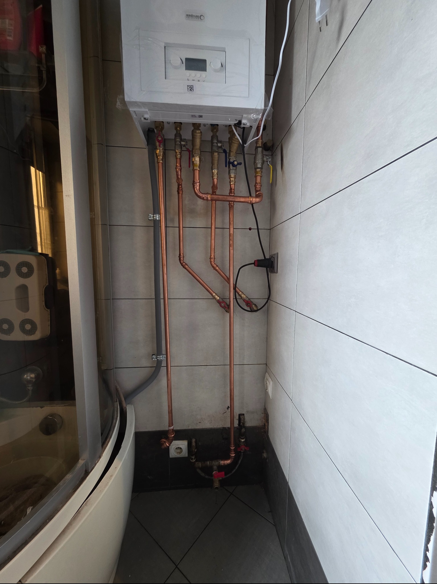 Nowy, biały piec gazowy zamontowany na ścianie z szarymi i białymi płytkami. Widoczne miedziane rury instalacji, podłączenia i elementy armatury. Obok kabina prysznicowa.