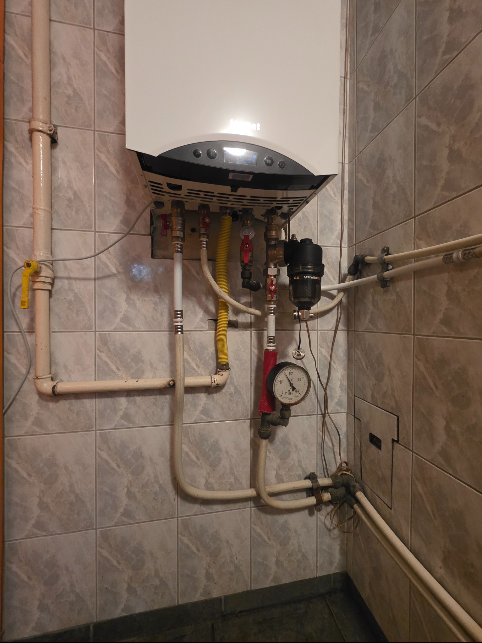 Biały piec gazowy z podłączeniami (żółty przewód gazowy, rury) na tle płytek. Widoczny manometr i filtr. Instalacja w pomieszczeniu.