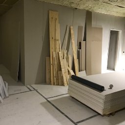 PITSERWIS - Wnętrze w trakcie prac wykończeniowych: surowe ściany z płyt kartonowo-gipsowych, stos płyt KG na podłodze z widocznymi liniami podziału, ułożone drewniane deski i wełna mineralna na suficie...