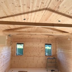 PITSERWIS - Wnętrze domu w trakcie wykańczania, ściany i sufit wyłożone jasnym drewnem, widoczne okna, drabina i narzędzia, podłoga częściowo zabezpieczona folią.