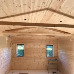 Wnętrze domu w trakcie wykańczania, ściany i sufit wyłożone jasnym drewnem, widoczne okna, drabina i narzędzia, podłoga częściowo zabezpieczona folią.