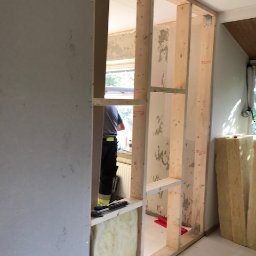 PITSERWIS - Wnętrze w trakcie tworzenia ścianki działowej: drewniany szkielet konstrukcji, częściowo obłożony płytą, widoczna wełna mineralna wewnątrz, pracownik w tle przy oknie.