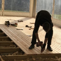 PITSERWIS - Pracownik w czarnym kombinezonie przykręca deski podłogowe w jasnym pomieszczeniu z przeszklonymi ścianami, widoczne narzędzia i surowy szkielet podłogi.