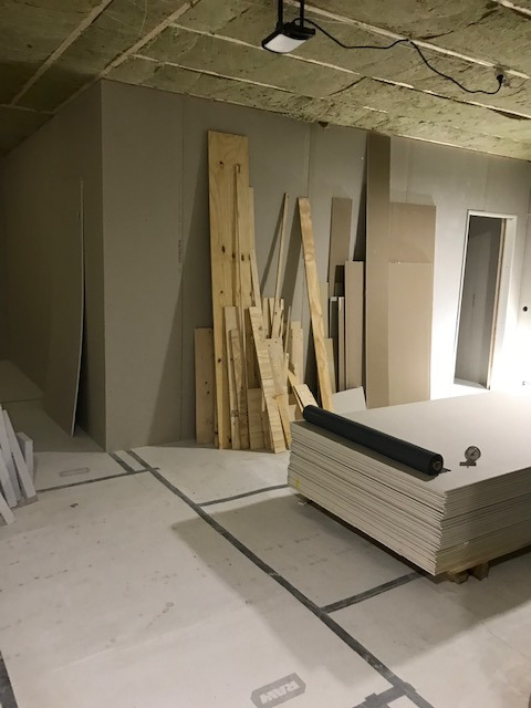 Wnętrze w trakcie prac wykończeniowych: surowe ściany z płyt kartonowo-gipsowych, stos płyt KG na podłodze z widocznymi liniami podziału, ułożone drewniane deski i wełna mineralna na suficie...