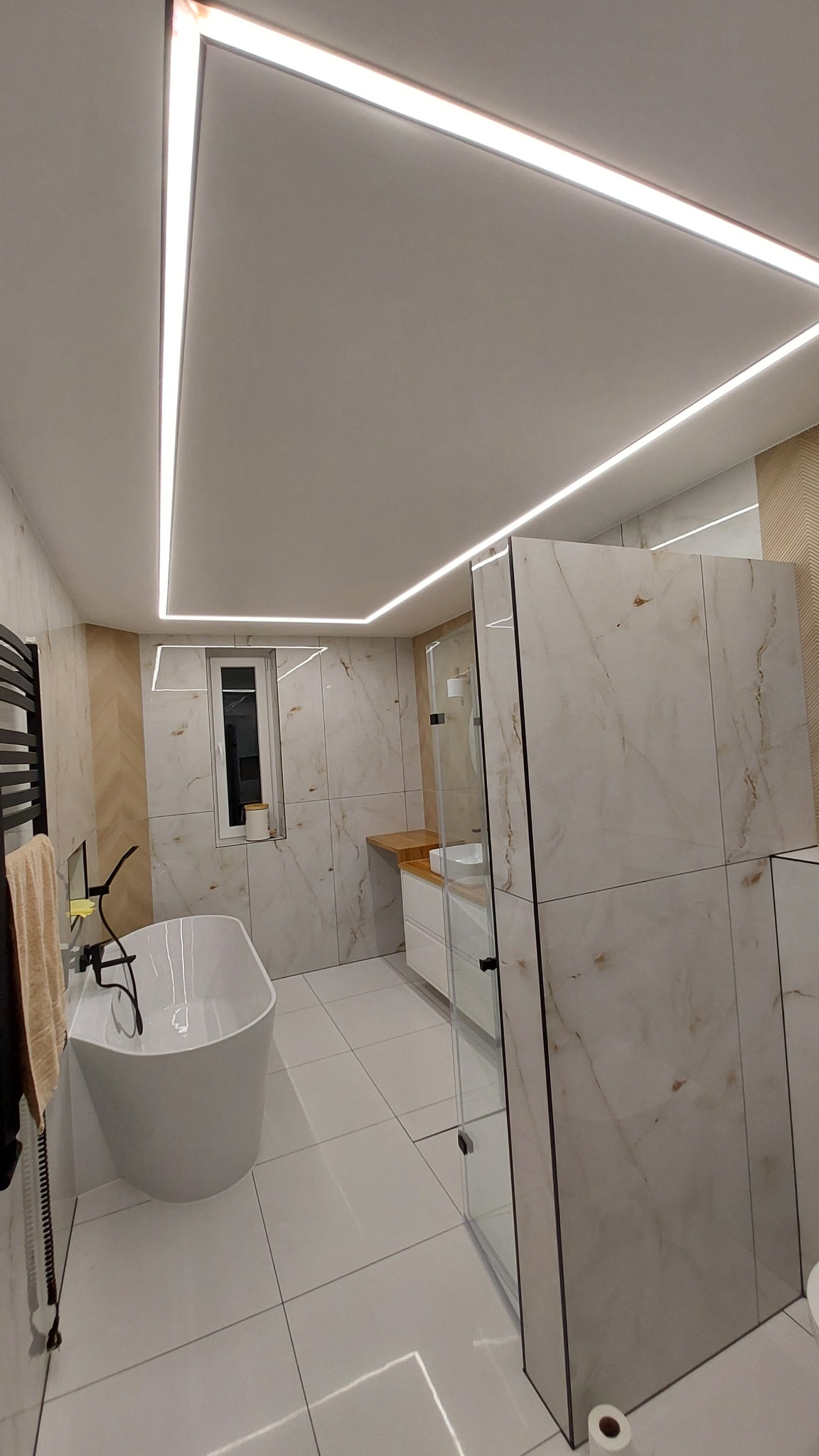 Nowoczesna łazienka z wolnostojącą wanną, kabiną prysznicową z marmurowymi płytkami i podświetleniem LED na suficie.