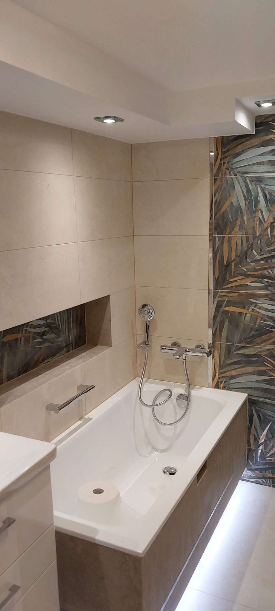 Nowoczesna łazienka po remoncie: beżowe płytki, wanna z hydromasażem, dekoracyjna ściana z motywem roślinnym, podświetlenie LED pod wanną i wnęka z ozdobnym panelem.