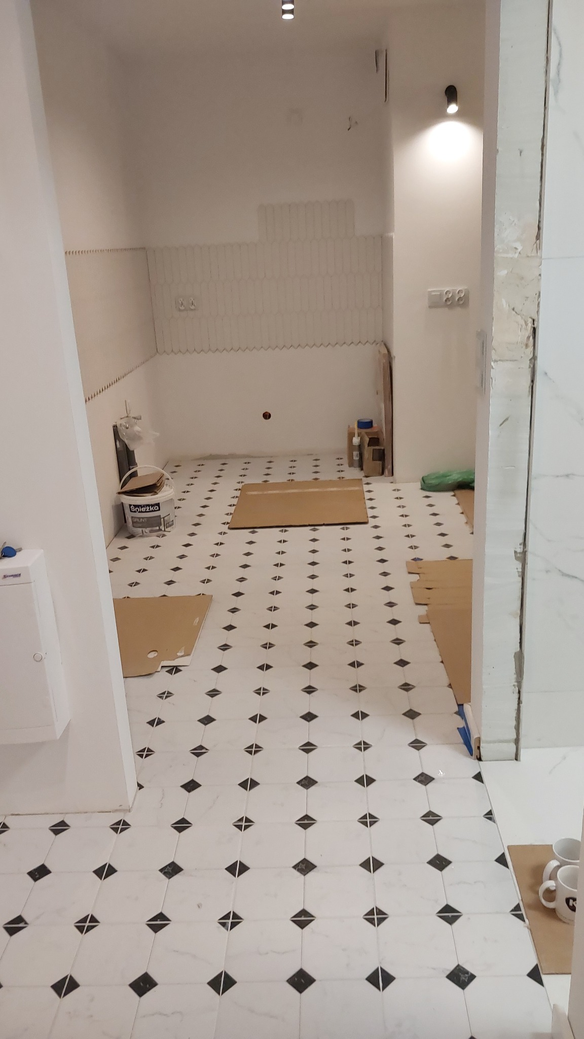 Wnętrze w trakcie prac wykończeniowych: biało-czarna podłoga w karo, częściowo zakryta kartonami ochronnymi, ściany przygotowane pod płytki, widoczne narzędzia i materiały budowlane.