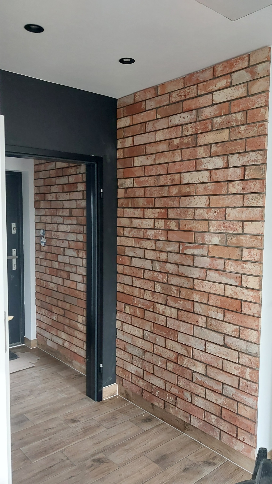 Wnętrze z dekoracyjną ścianą z cegły klinkierowej, brązowe panele podłogowe imitujące drewno, czarna rama drzwiowa i wpuszczane oświetlenie sufitowe.
