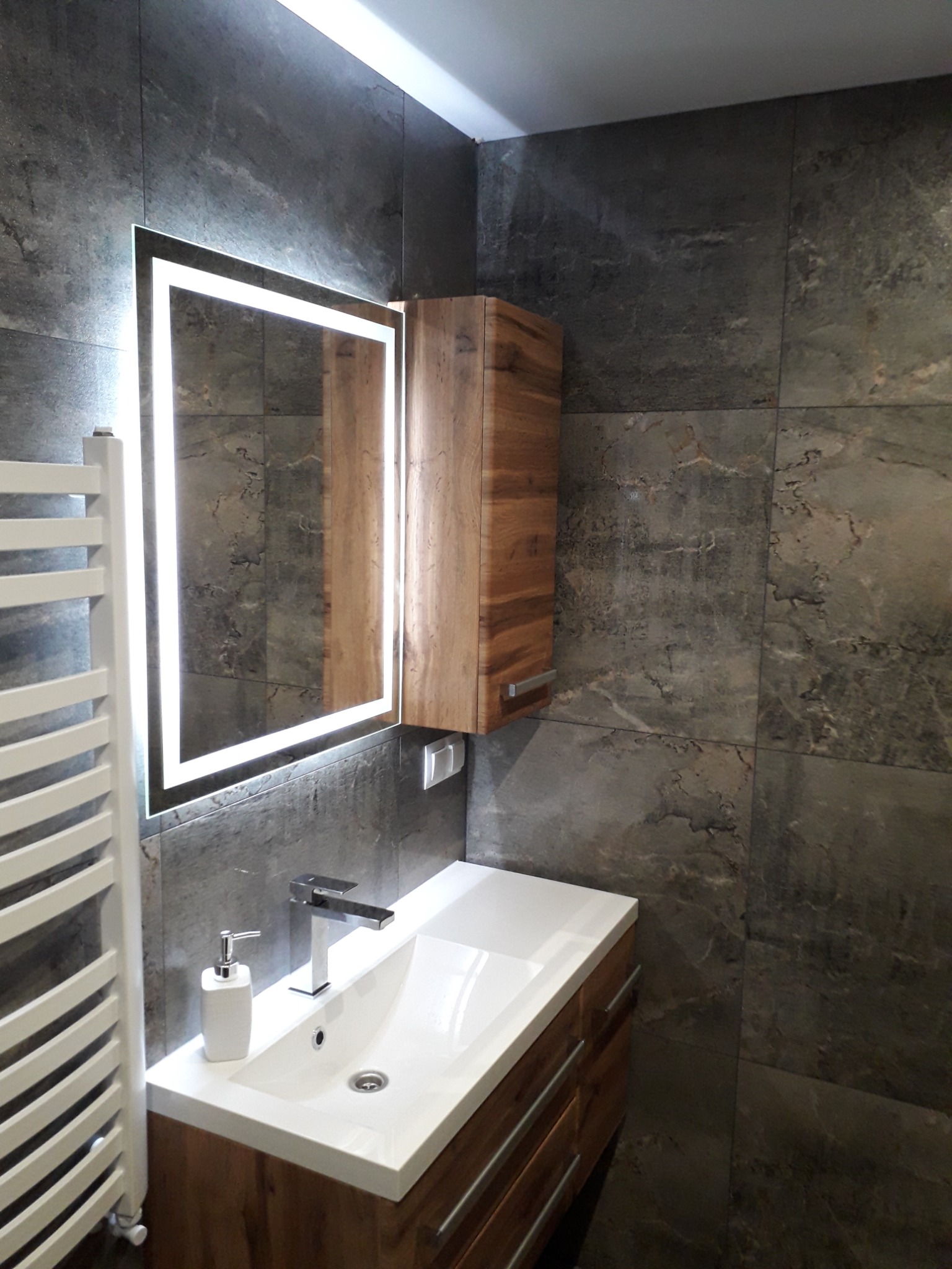Nowoczesna łazienka z szarymi płytkami imitującymi kamień, podświetlanym lustrem, drewnianą szafką pod umywalką i grzejnikiem drabinkowym.