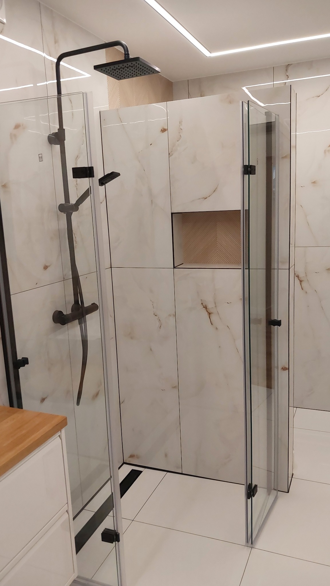 Nowoczesna kabina prysznicowa z czarną armaturą i płytkami imitującymi marmur, z wnęką wykończoną drewnem.