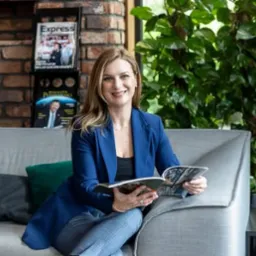 Uśmiechnięta kobieta w niebieskiej marynarce siedzi na szarej sofie, czytając magazyn; w tle regał z czasopismami i zielone rośliny.