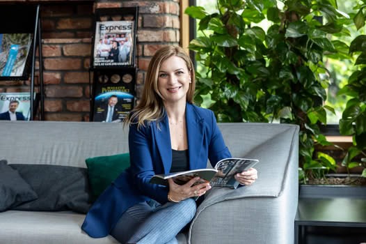 Uśmiechnięta kobieta w niebieskiej marynarce siedzi na szarej sofie, czytając magazyn; w tle regał z czasopismami i zielone rośliny.