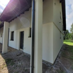 Firma Remontowo-Budowlana Daniel Olek - Świeżo otynkowana i pomalowana elewacja domu jednorodzinnego z widocznymi czarnymi rynnami i oknami, w tle drzewa i niezagospodarowany teren.