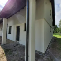Świeżo otynkowana i pomalowana elewacja domu jednorodzinnego z widocznymi czarnymi rynnami i oknami, w tle drzewa i niezagospodarowany teren.