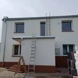 Dwukondygnacyjny dom po remoncie elewacji, z widocznymi drabinami, świeżo pomalowaną fasadą w odcieniach bieli i brązu, oraz stosem białych płyt izolacyjnych obok
