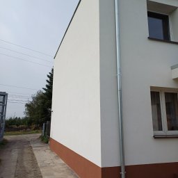 Firma Remontowo-Budowlana Daniel Olek - Świeżo otynkowana i pomalowana elewacja budynku z zamontowaną rynną spustową, widoczny cokół w kolorze brązowym oraz narzędzia na chodniku.
