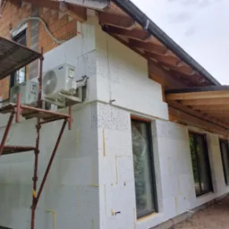 Dom w trakcie prac, z widocznym ociepleniem ścian styropianem grafitowym, rusztowaniem, zamontowanymi klimatyzatorami i elementami konstrukcji dachu.