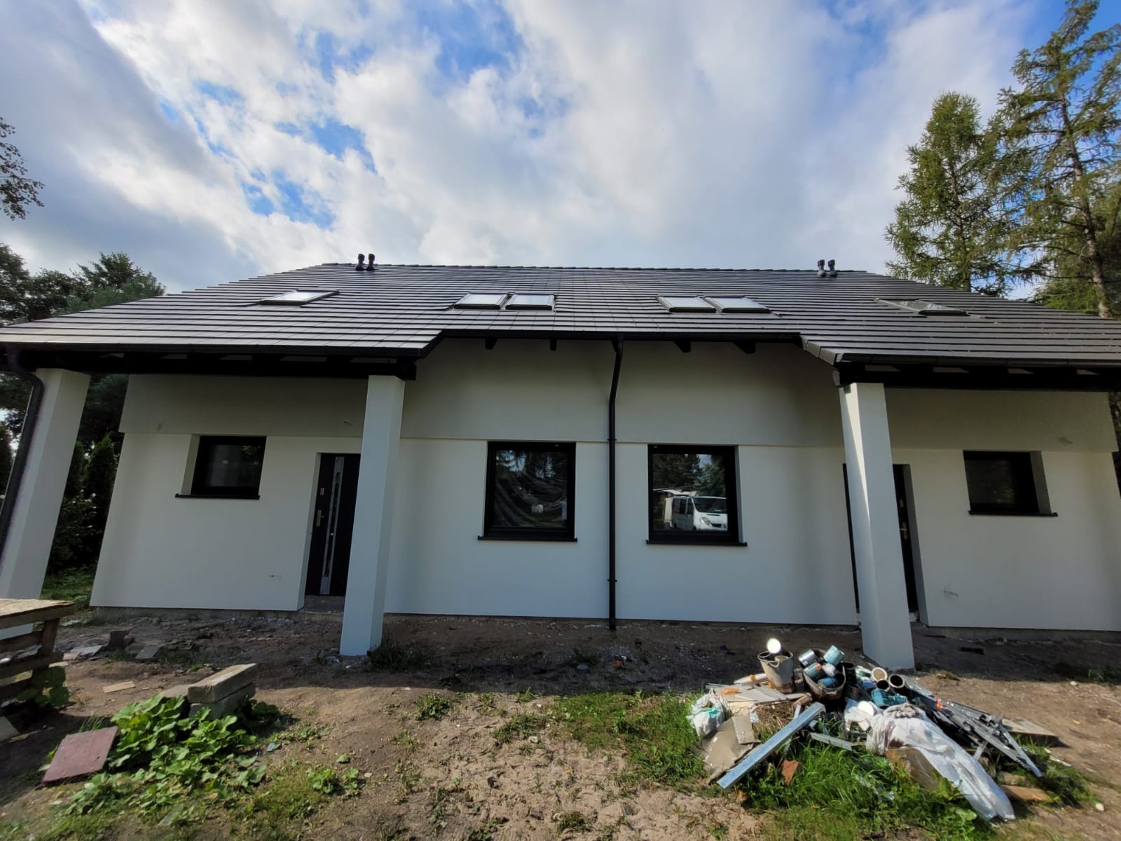 Świeżo otynkowany dom jednorodzinny z ciemną dachówką i oknami, widoczne ślady po pracach wykończeniowych na trawniku.