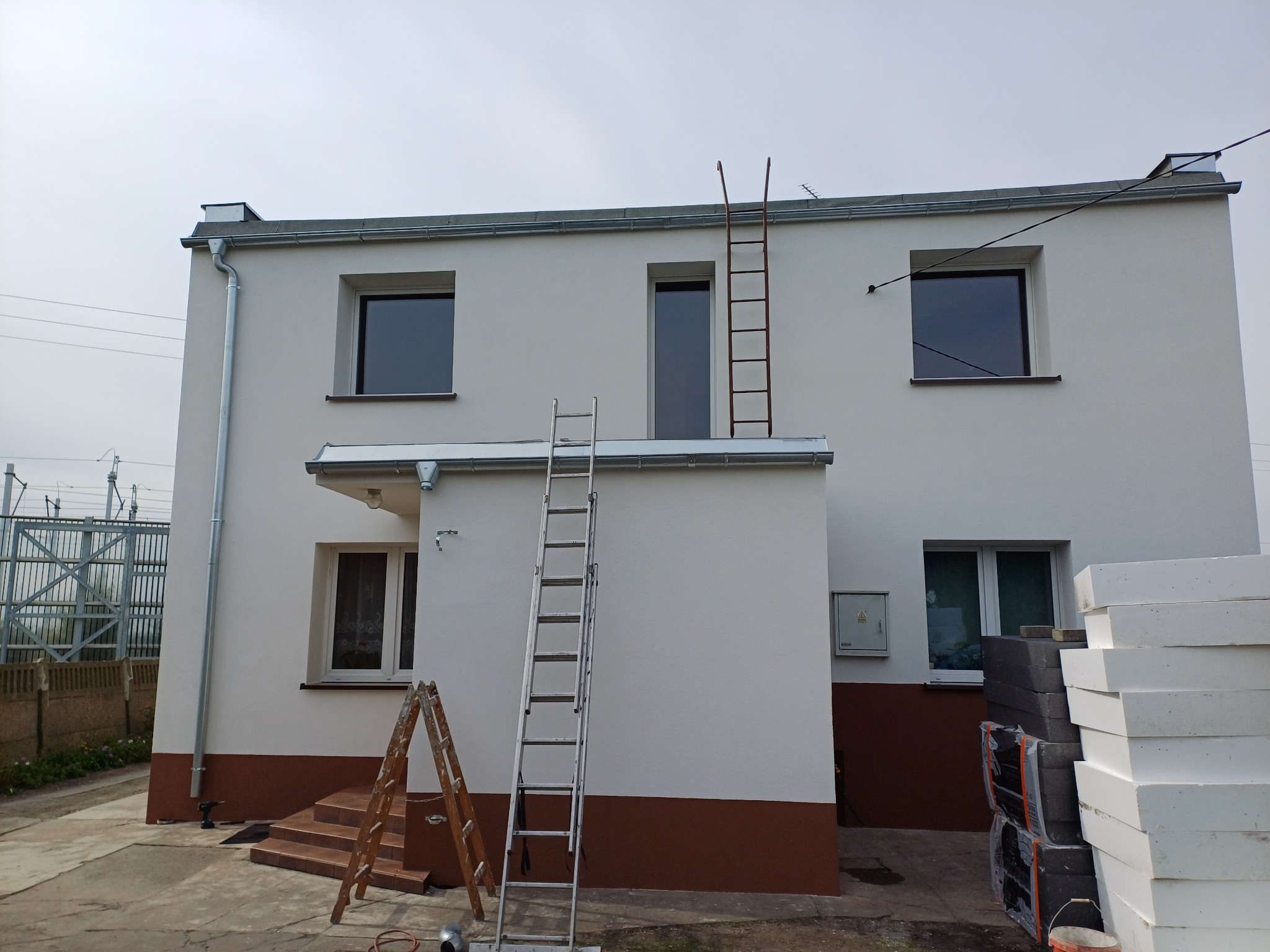 Dwukondygnacyjny dom po remoncie elewacji, z widocznymi drabinami, świeżo pomalowaną fasadą w odcieniach bieli i brązu, oraz stosem białych płyt izolacyjnych obok