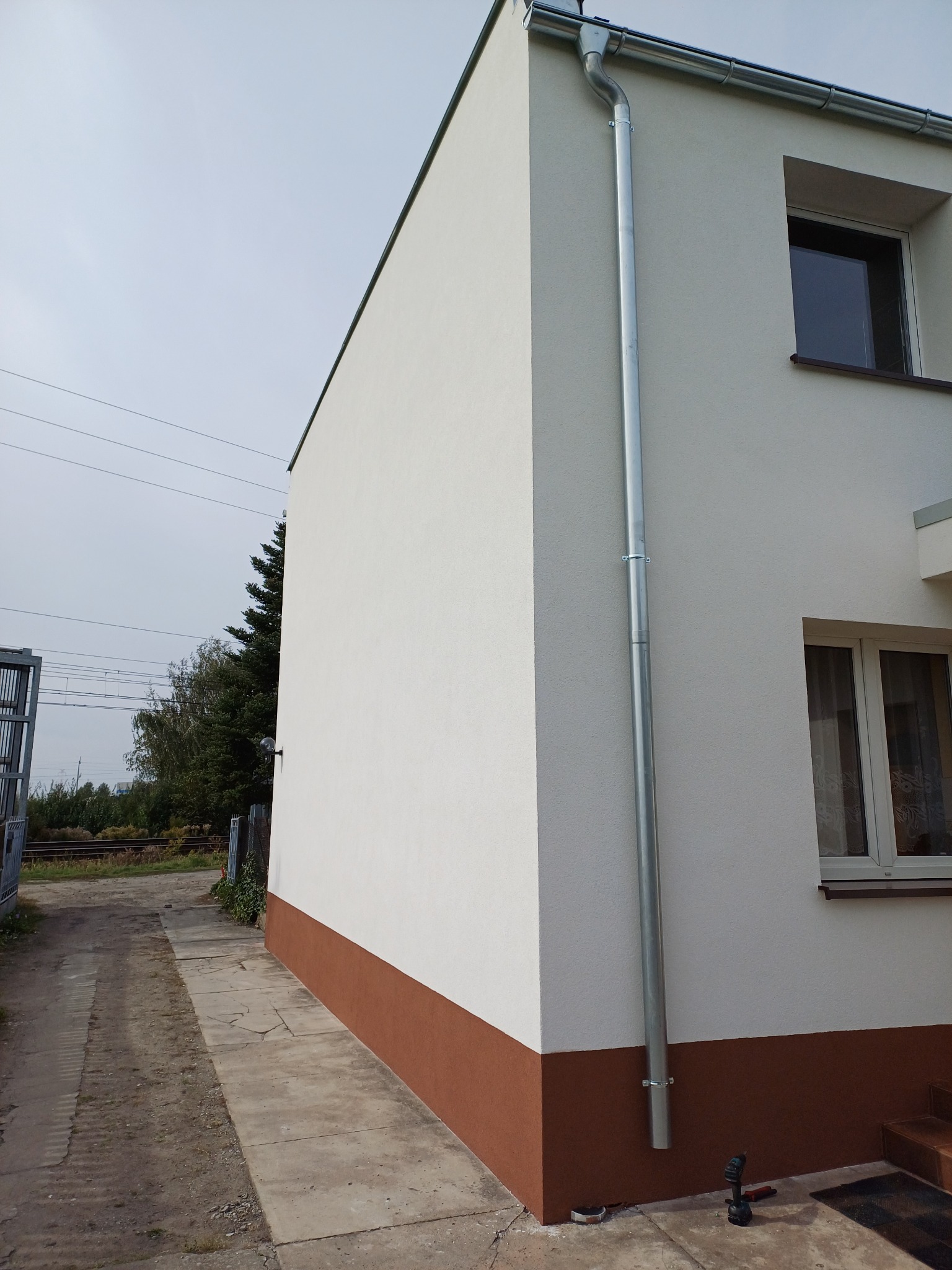 Świeżo otynkowana i pomalowana elewacja budynku z zamontowaną rynną spustową, widoczny cokół w kolorze brązowym oraz narzędzia na chodniku.