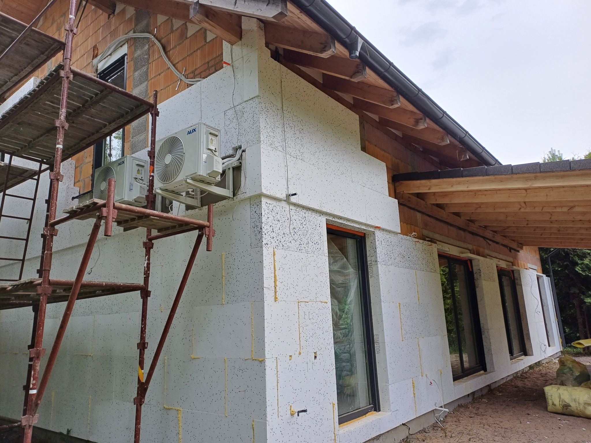 Dom w trakcie prac, z widocznym ociepleniem ścian styropianem grafitowym, rusztowaniem, zamontowanymi klimatyzatorami i elementami konstrukcji dachu.