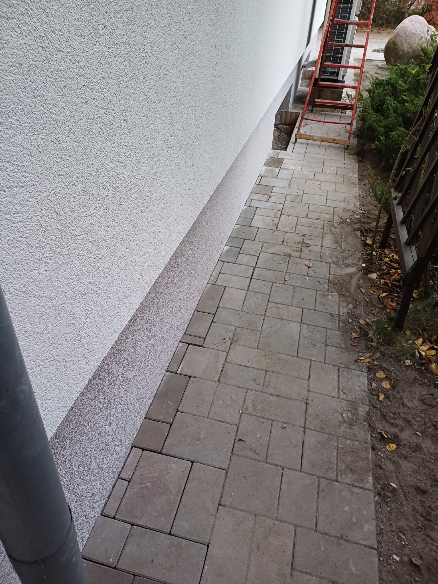 Ściana budynku z fakturą baranka, cokół z ciemniejszego tynku, obok ułożona kostka brukowa, widoczna czerwona drabina oparta o ścianę.