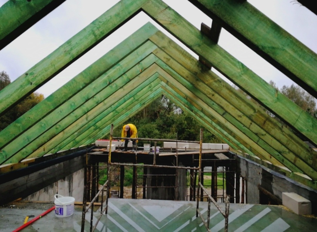 Konstrukcja dachu z zielonych belek widziana od wewnątrz, pracownik w żółtej kurtce na rusztowaniu, widoczne narzędzia i wiadro z farbą.