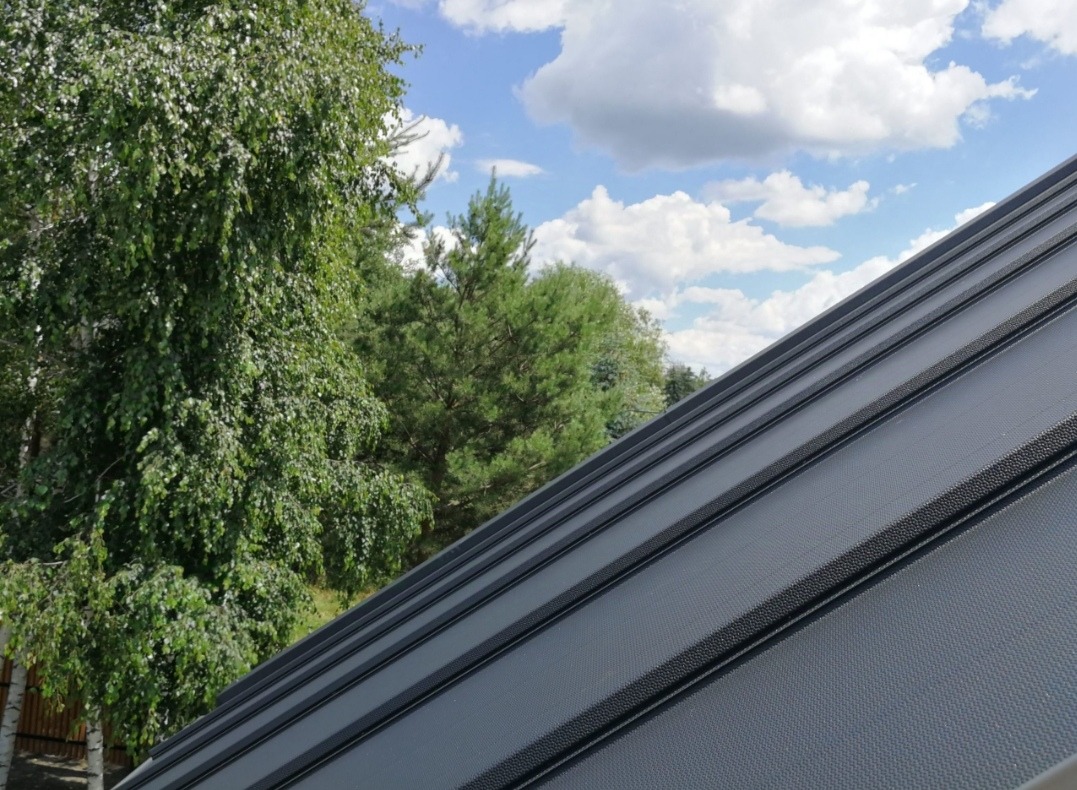 Szary, blaszany dach trapezowy z widocznym łączeniem paneli, na tle błękitnego nieba z chmurami i zieleni drzew.