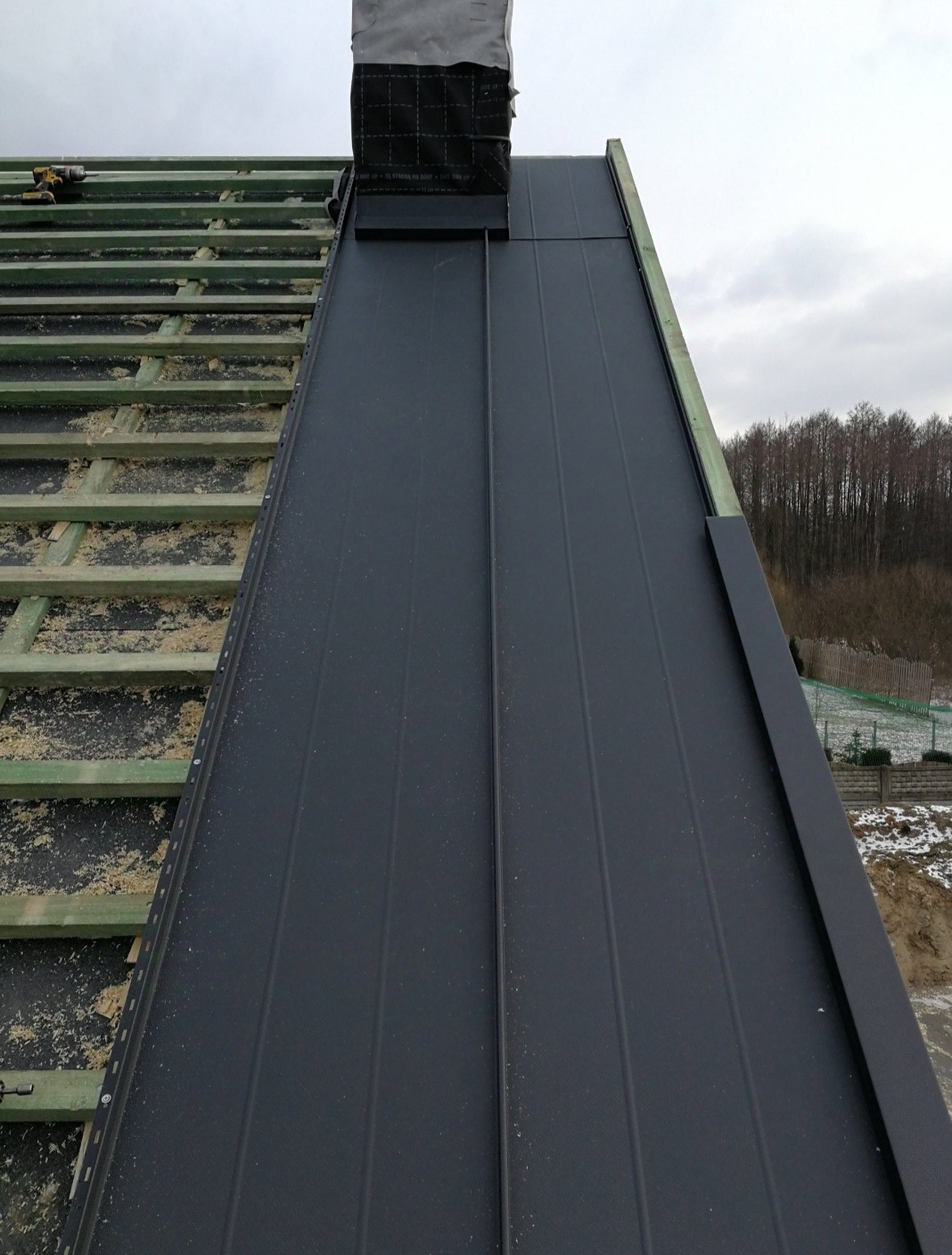 Montaż paneli na dachu z widoczną konstrukcją więźby i kominem zabezpieczonym membraną, w tle drzewa i fragment krajobrazu.