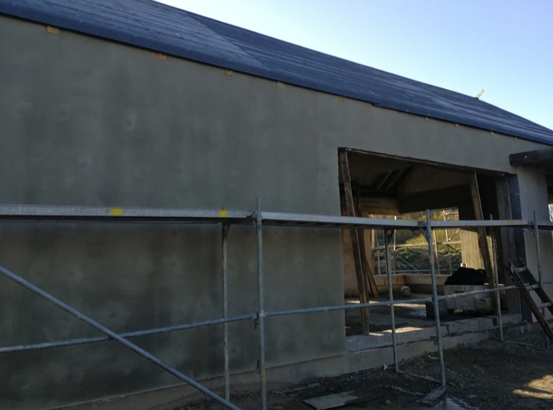 Świeżo otynkowana ściana zewnętrzna budynku z widocznym fragmentem dachu, rusztowaniem i surową konstrukcją okienną, w tle zieleń.