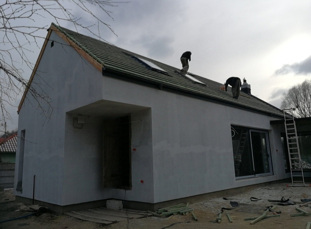 Dwóch dekarzy pracuje na dachu nowo wybudowanego, otynkowanego domu jednorodzinnego z oknami dachowymi, widoczne resztki materiałów budowlanych na ziemi.