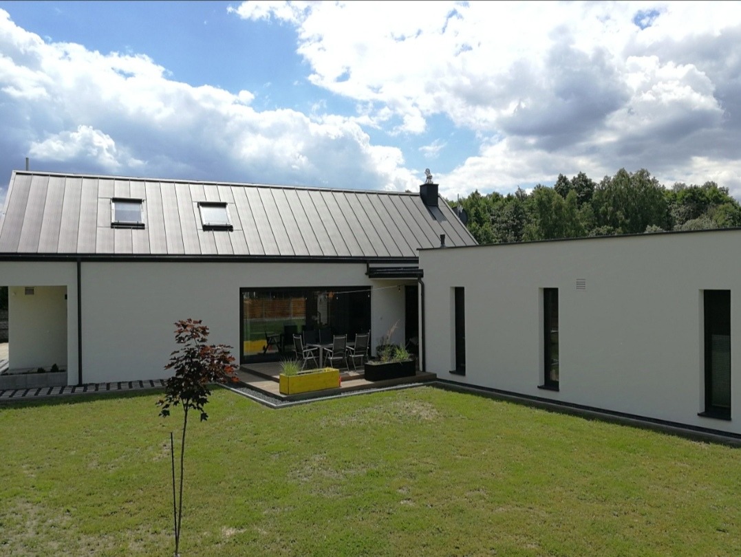 Nowoczesny dom z grafitowym dachem blaszanym na rąbek stojący, białymi ścianami i dużymi oknami, widok z ogrodu w słoneczny dzień.