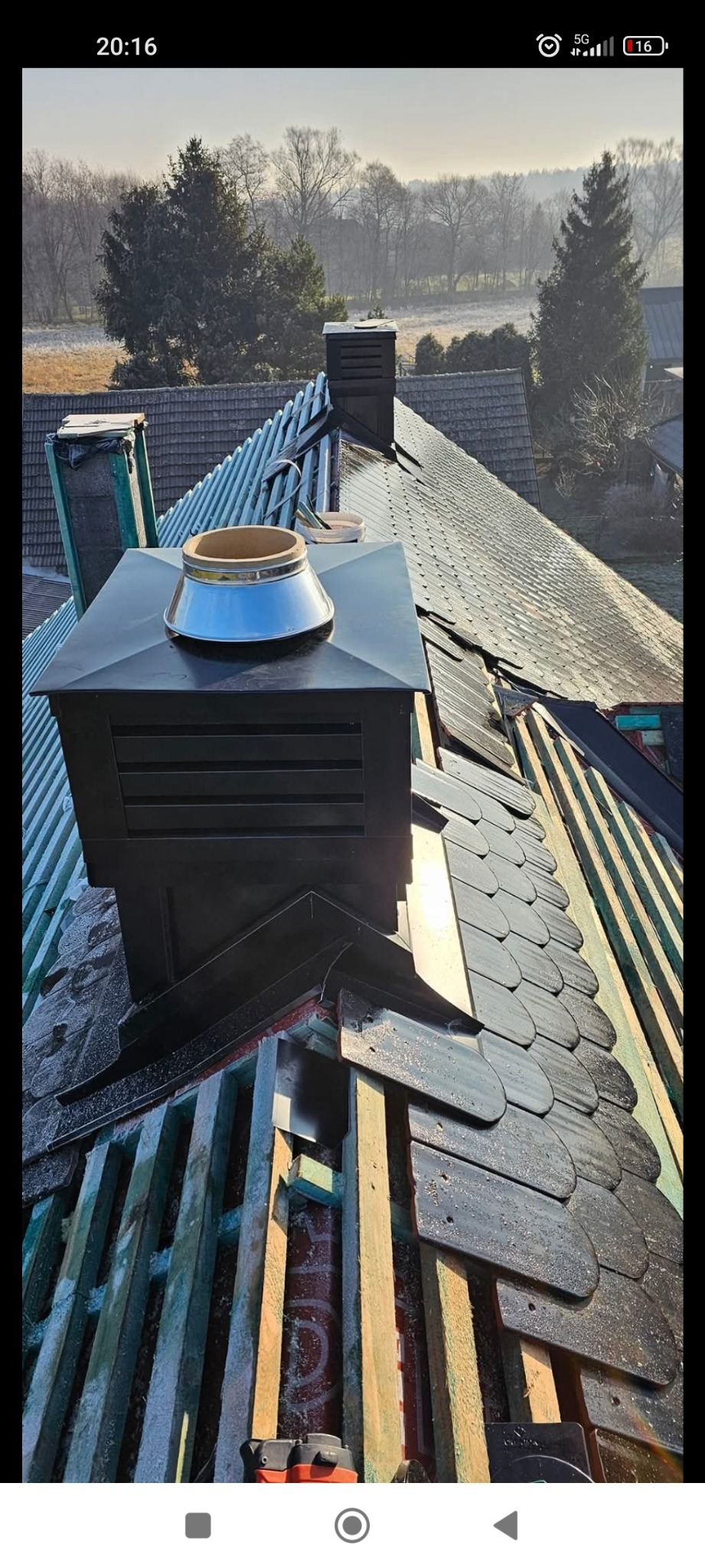 Montaż czarnego komina z wentylacją na dachu krytym łupkiem, widoczna konstrukcja drewniana i ocieplenie, w tle zalesiony krajobraz w porannym słońcu.