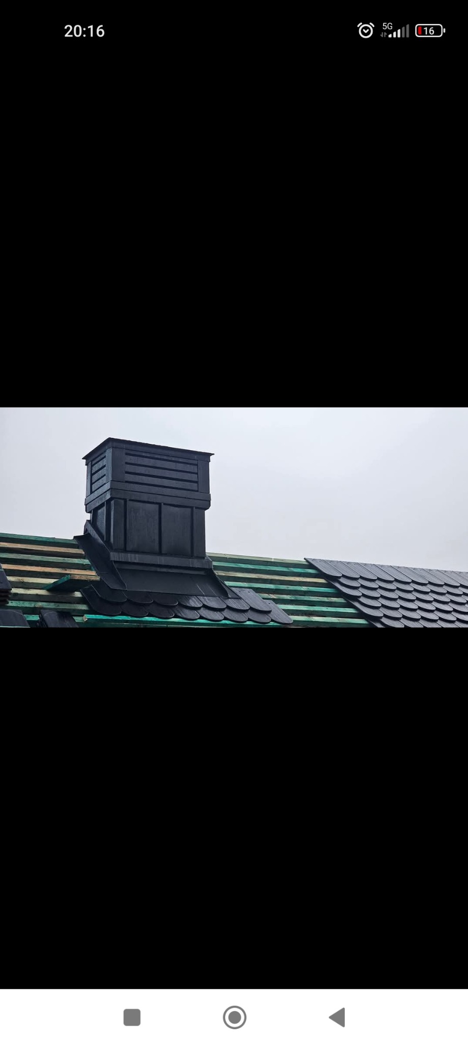 Czarny komin z nową obróbką blacharską i częściowo ułożonym dachem z dachówki bitumicznej na tle pochmurnego nieba.