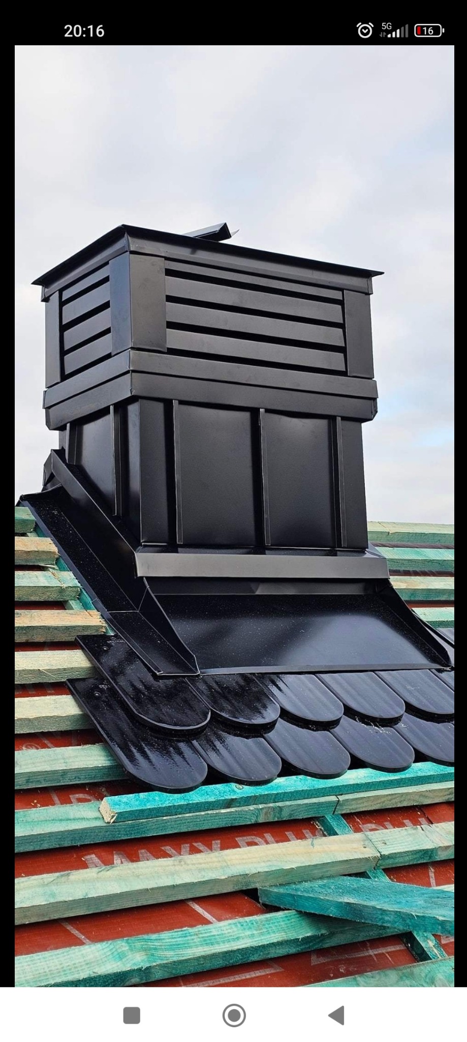 Czarny, metalowy komin z obróbką blacharską na dachu pokrytym dachówką karpiówką, widoczne elementy drewnianej konstrukcji dachu.