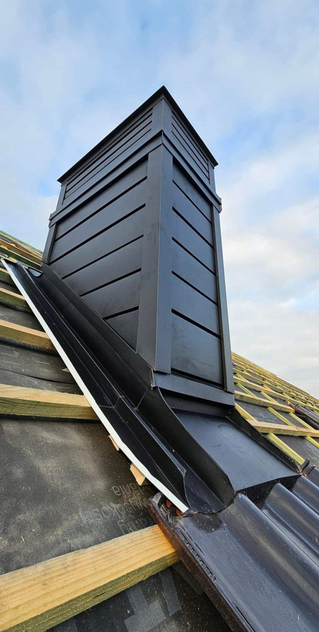 Nowoczesny, czarny komin z obróbką blacharską na dachu w trakcie budowy, widoczne elementy więźby dachowej i dachówki ceramicznej.
