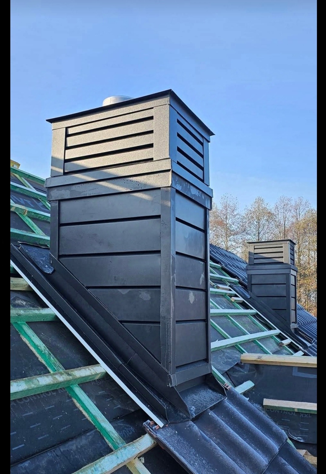 Nowoczesny, czarny komin z blachy na dachu w trakcie budowy, widoczna konstrukcja więźby dachowej i membrana, drugi komin w tle.