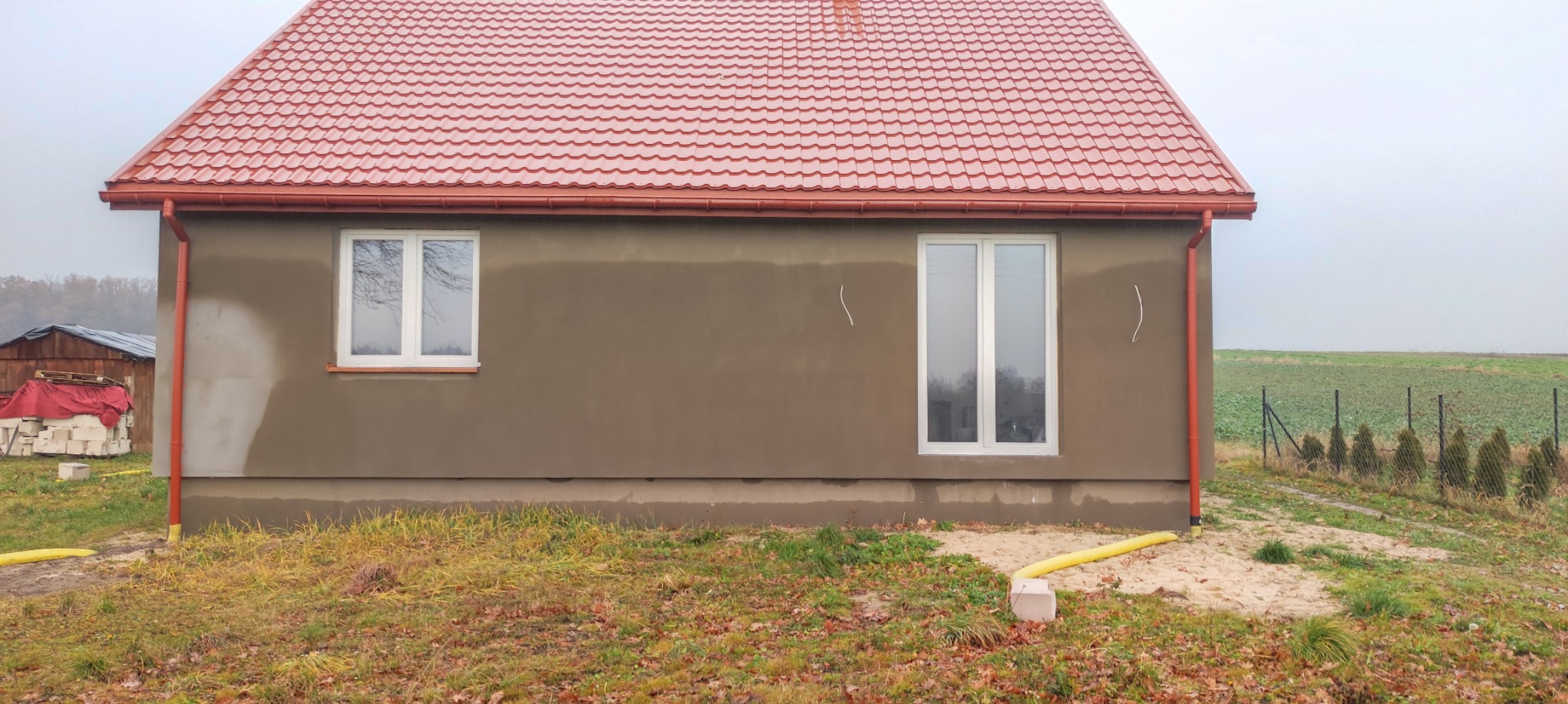 Dom jednorodzinny z nową, czerwoną dachówką, otynkowaną elewacją w trakcie prac wykończeniowych, widoczne ślady po łatach, rynny w kolorze dachu, trawnik z opadłymi liśćmi.