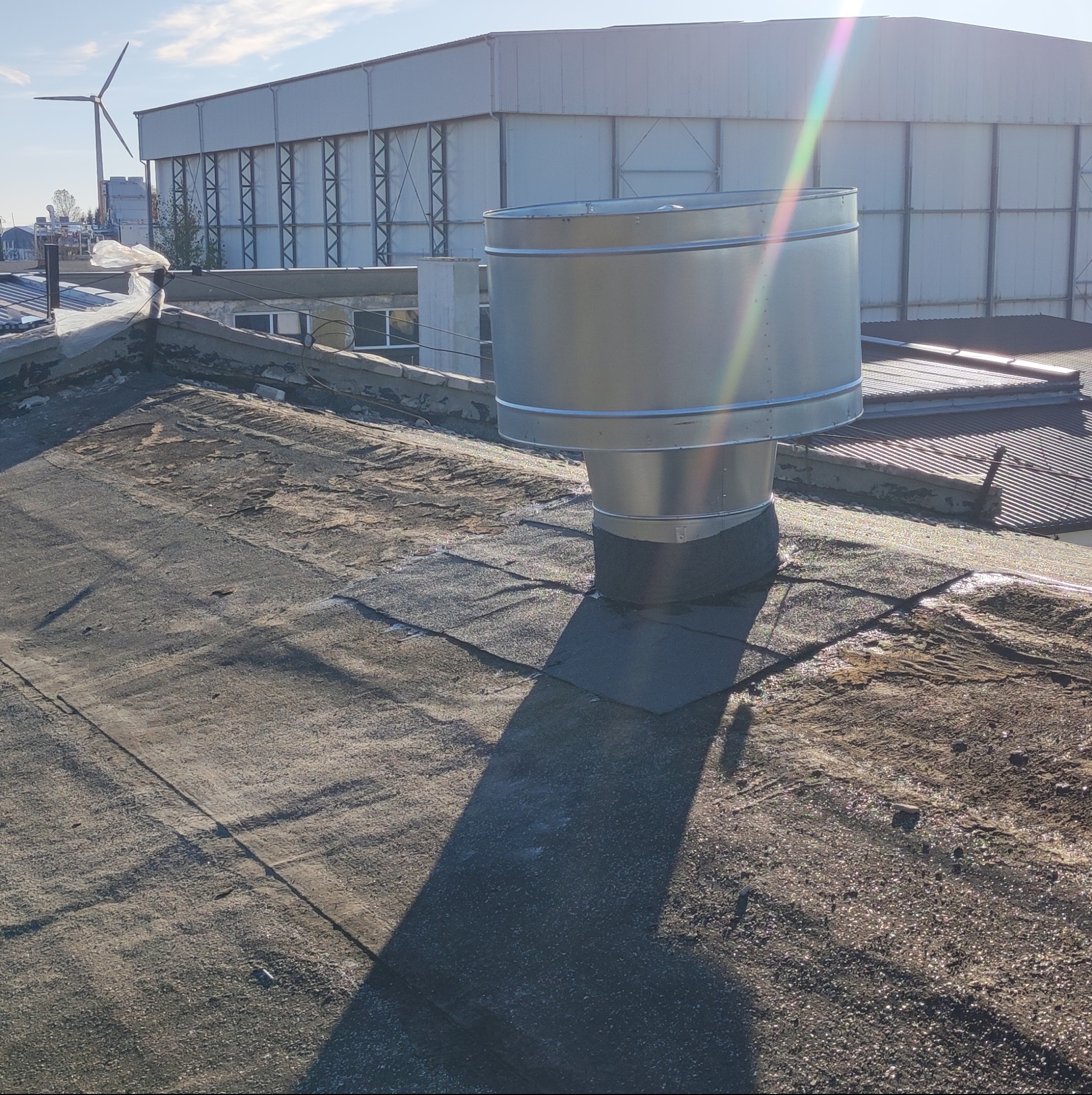 Metalowy komin wentylacyjny na płaskim, bitumicznym dachu budynku przemysłowego; w tle widoczny budynek magazynowy i turbina wiatrowa.