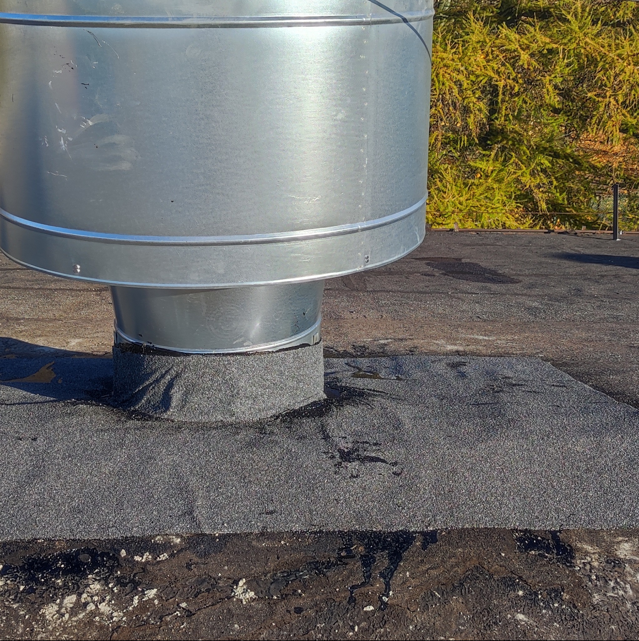 Metalowy komin wentylacyjny na dachu pokrytym papą, zabezpieczony kołnierzem uszczelniającym; w tle zielone drzewo iglaste.