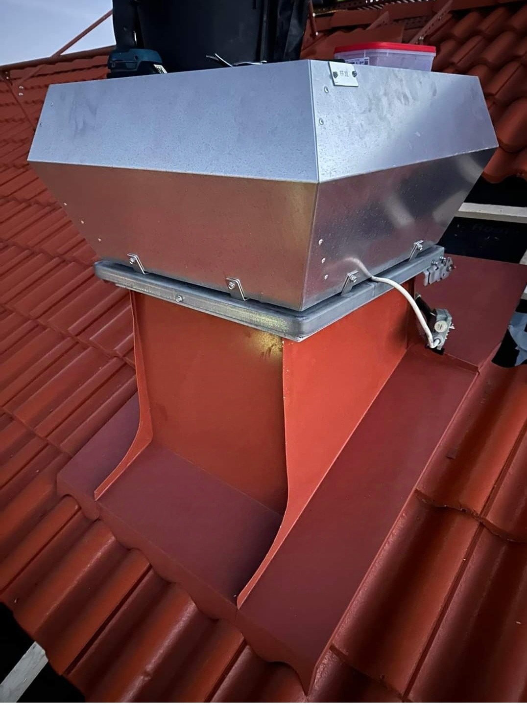 Nowa, błyszcząca obróbka blacharska komina wentylacyjnego na dachu z czerwonej dachówki ceramicznej, widoczne mocowania i przewód elektryczny.