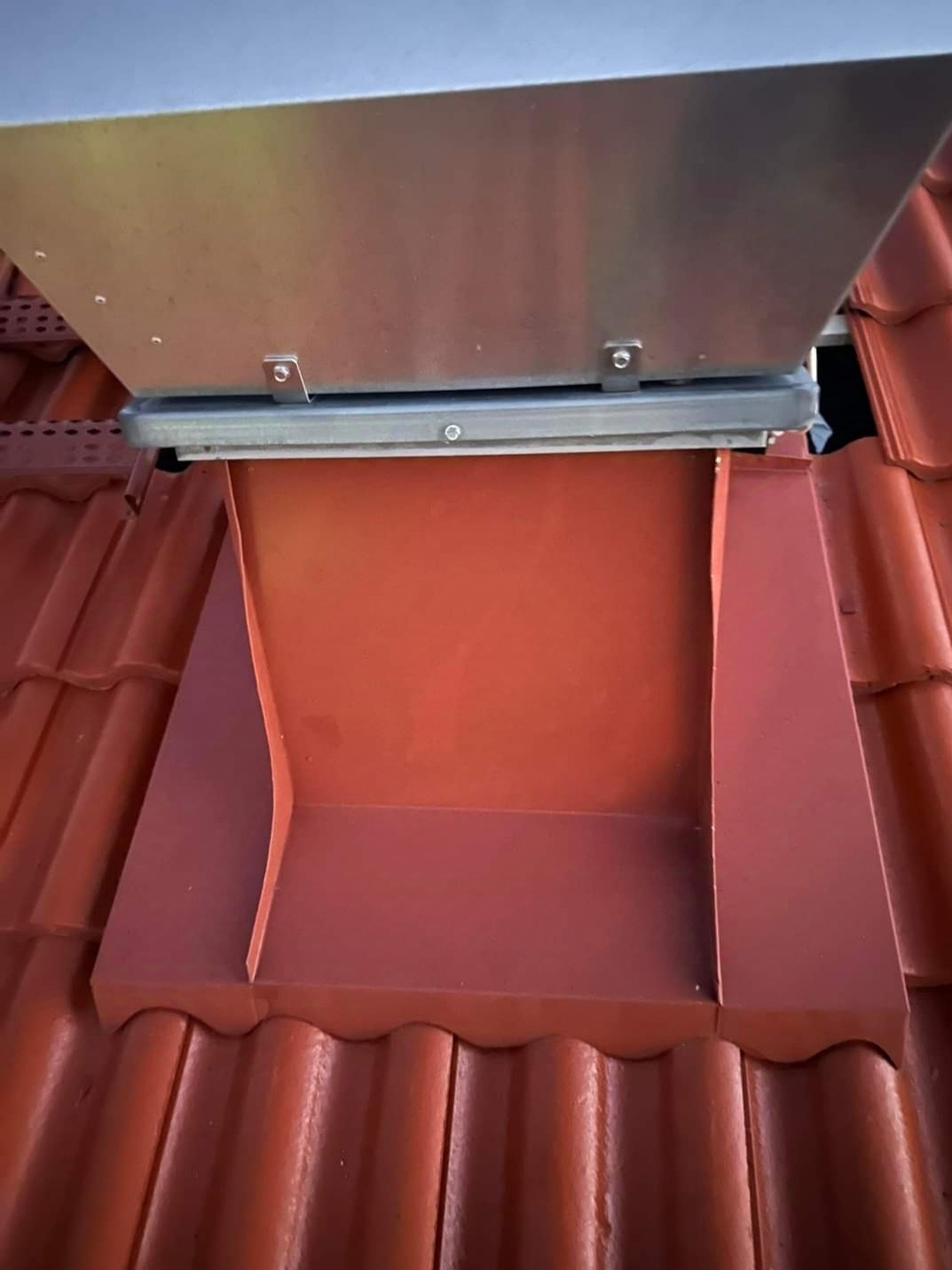 Czerwona blaszana obróbka komina na dachu z dachówki ceramicznej, widoczne mocowania i elementy konstrukcyjne.