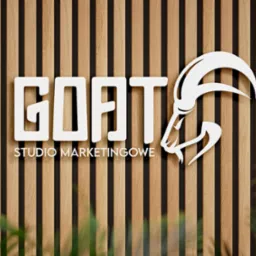 Logo 'GOAT Studio Marketingowe' w kolorze białym, umieszczone na tle z drewnianych listew. Litera 'T' zastąpiona stylizowaną głową kozła z zakręconymi rogami. Częściowo widoczne zielone rośliny...