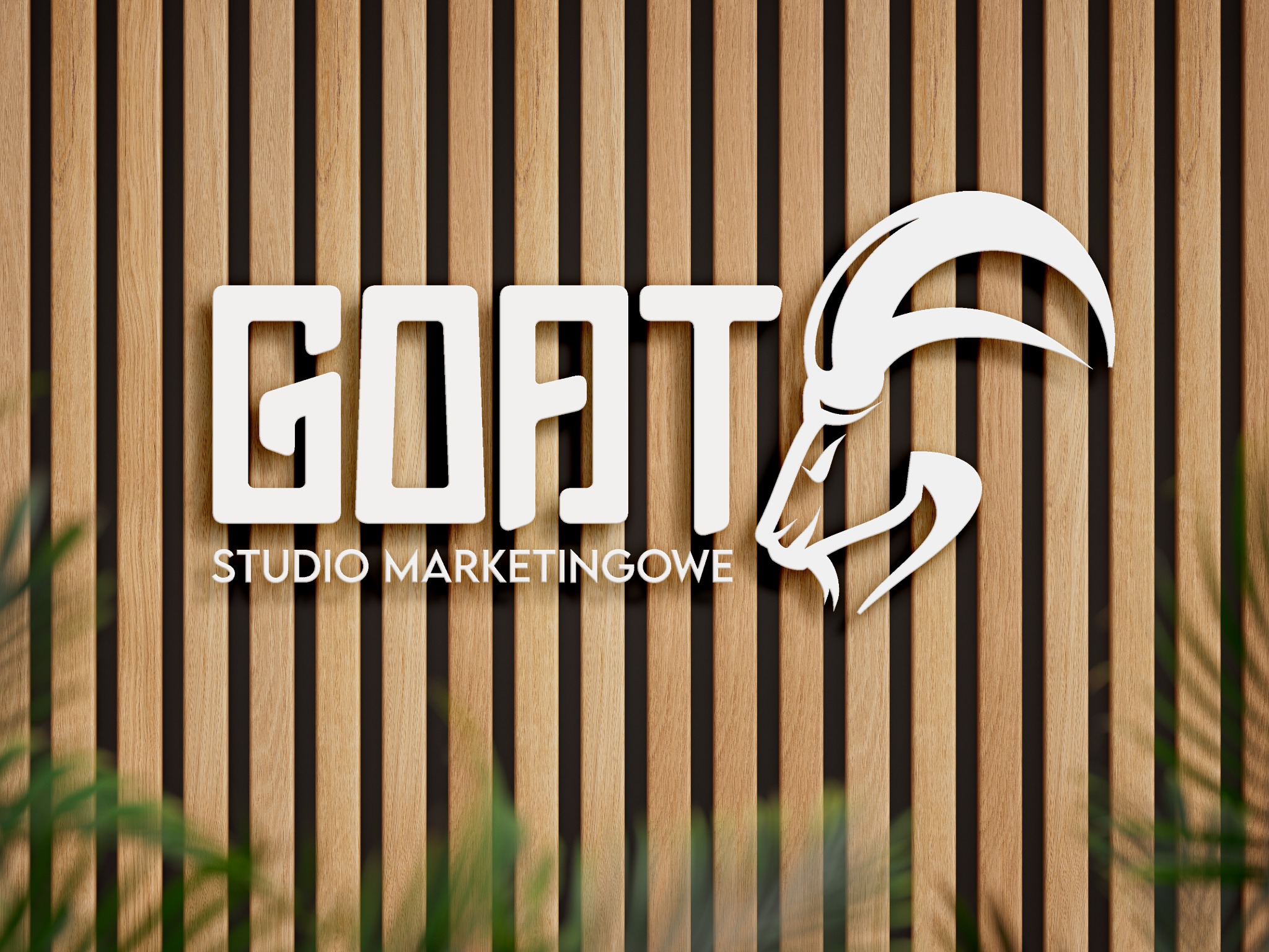 Logo 'GOAT Studio Marketingowe' w kolorze białym, umieszczone na tle z drewnianych listew. Litera 'T' zastąpiona stylizowaną głową kozła z zakręconymi rogami. Częściowo widoczne zielone rośliny...
